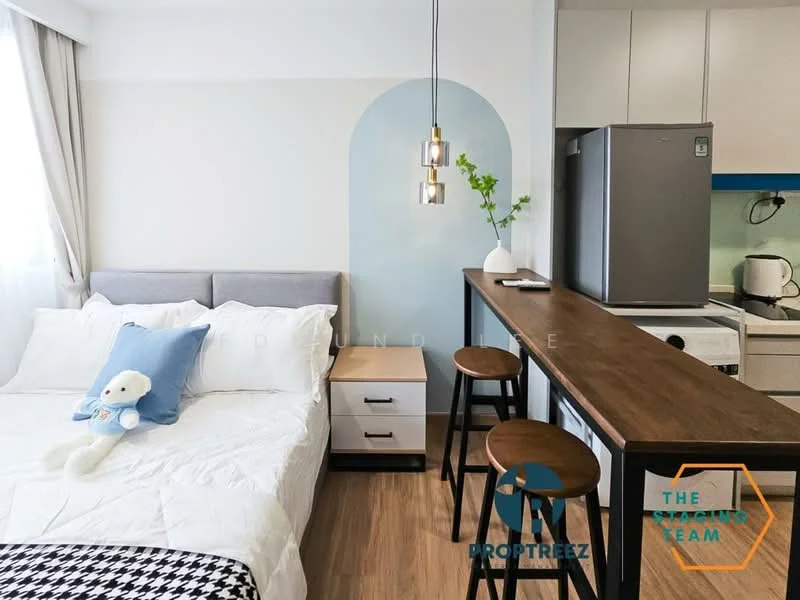 Room features: queen-size bed with white bedding, grey upholstered headboard, nightstand with drawer, wooden bar counter, two wooden stools, refrigerator, kitchen cabinetry in blue, white walls, light wood flooring, pendant light fixture, decorative plant on counter, window with sheer curtains; style: modern.