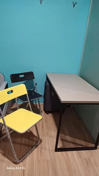 Room features: light blue painted walls, wood laminate flooring, folding black chair, folding yellow chair, simple rectangular table with light-colored top and black metal legs, luggage bag, wall hooks; style: modern.