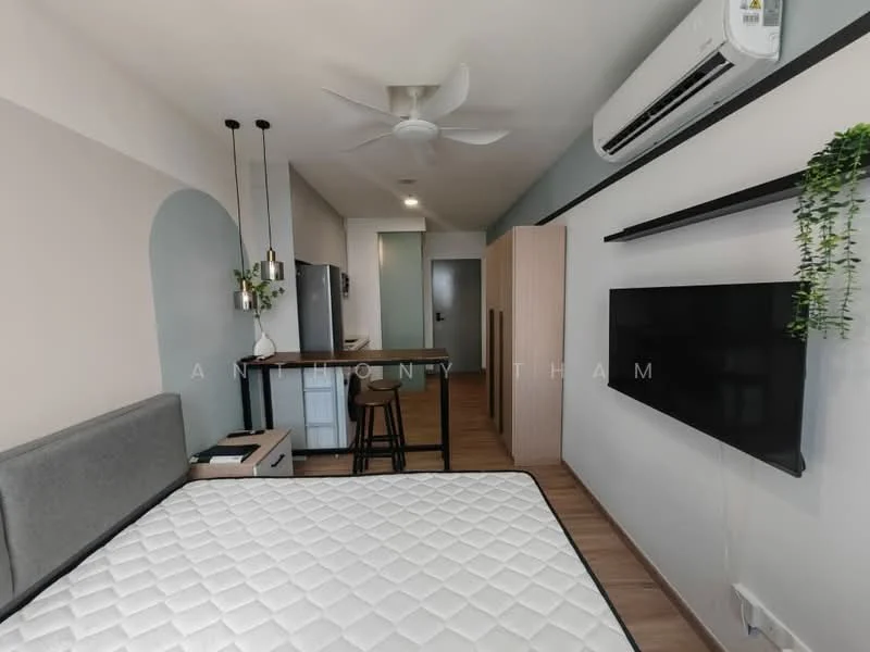 Room features: queen-size bed, upholstered headboard, wall-mounted TV, wooden flooring, 5-blade ceiling fan with light, air conditioning unit, built-in wardrobe, kitchenette with bar counter, stool seating at bar, refrigerator, wall-mounted shelving, decorative plant, light-colored walls, open layout; style: modern.