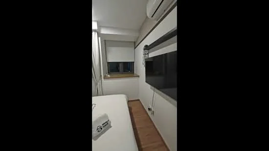 Room features: white painted walls, wooden flooring, floor-to-ceiling window, roller blinds, wall-mounted TV, air conditioning unit, bedside table, decorative wall panel, light fixture; style: modern.