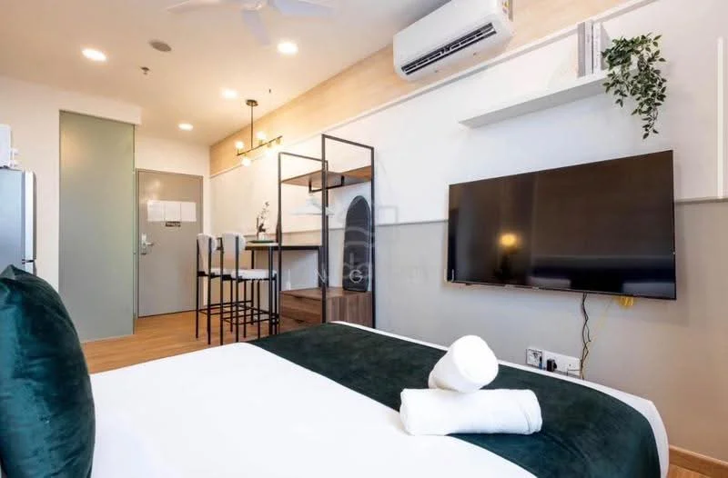 Room features: queen-size bed with green accent, white bedding, wall-mounted TV, wooden flooring, 1 wall-mounted split AC, 5-blade ceiling fan, open shelving unit, small dining table with 2 stools, refrigerator, decorative plant, light fixtures, painted walls with neutral tones, door to hallway; style: modern.