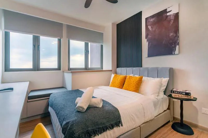 Bedroom features: queen-size bed with grey upholstered headboard, white bedding with decorative pillows in yellow, wooden flooring, two windows with roller blinds, wall-mounted artwork with abstract design, built-in desk with storage, ceiling fan, accent wall with vertical wood slats, bedside table, towel rolled on the bed; style: modern; view: city.