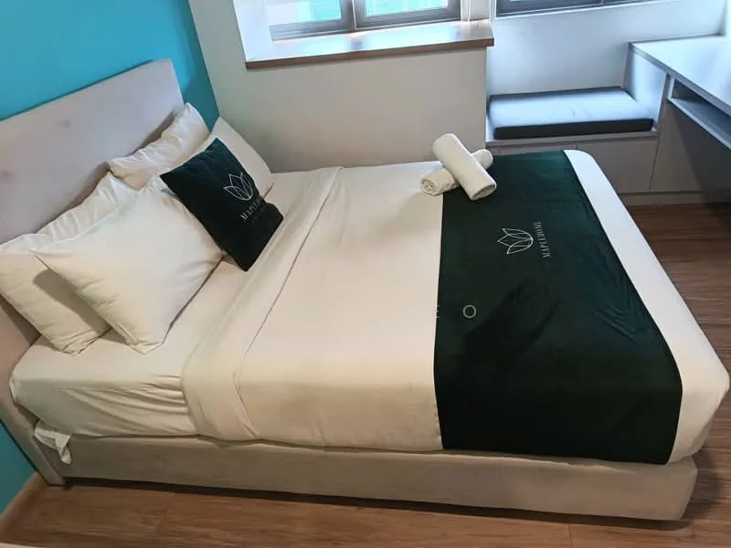 Bedroom features: queen-size bed, upholstered headboard, white bedding, green throw blanket, two white pillows, rolled white towels, wooden flooring, blue accent wall, window with natural light, built-in storage unit, window seat; style: modern.