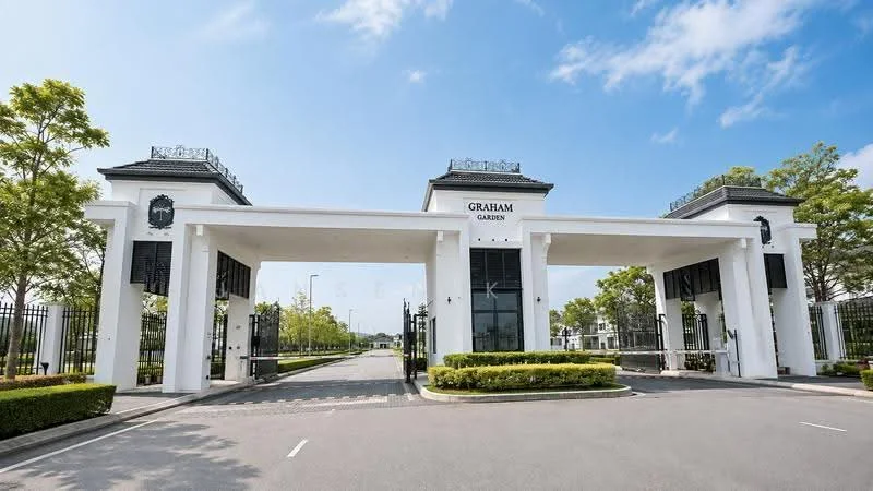 Exterior type: main entrance gate; features: white painted facade, two large entrance arches, black wrought iron gates, landscaped greenery, manicured hedges, paved driveway, decorative roof elements, signage with 'Graham Garden', surrounding trees, blue sky with clouds; style: modern.