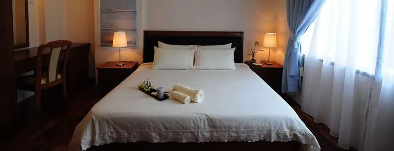 Bedroom features: king-size bed with white bedding, upholstered headboard, two bedside lamps with warm light, wooden bedside tables, wooden flooring, dressing table with mirror, chair at dressing table, floor-to-ceiling windows with sheer curtains, decorative plant on bed, towels rolled on bed; style: modern.