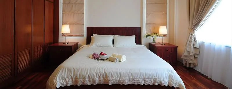 Bedroom features: king-size bed with white bedding, wooden headboard, two bedside lamps, wooden nightstands, decorative tray with fruits, white curtains, wooden flooring, built-in wardrobe with wooden panels, wall-mounted artwork, draped window treatment; style: modern.