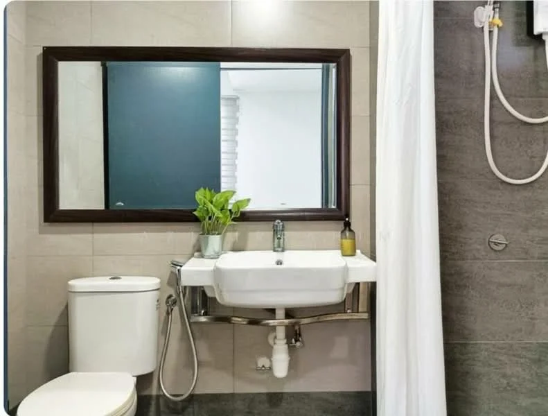 Bathroom features: white ceramic toilet, single-sink vanity with wall-mounted basin, chrome basin mixer faucet, large framed mirror, grey tile flooring, grey wall tiles, white shower curtain, potted plant on vanity, wall-mounted shower head, soap dispenser, towel rack; style: modern.