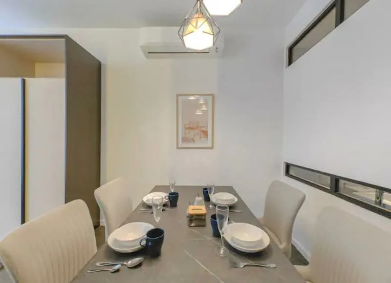 Dining features: rectangular dining table, 4 upholstered dining chairs, white plates, blue cups, cutlery, decorative centerpiece, wall-mounted air conditioning unit, framed artwork on wall, large window with sheer curtains, light fixture with geometric design; style: modern.