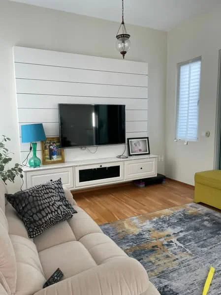 Living features: beige fabric sofa, coffee table, wall-mounted TV, TV console with storage, decorative framed artwork, green table lamp, area rug with abstract pattern, wood flooring, white accent wall with horizontal panels, window with blinds, hanging pendant light; style: modern.