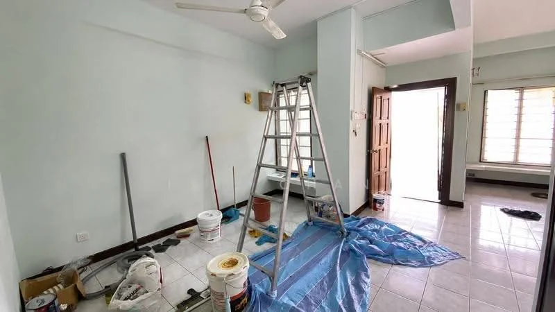 Room features: light blue painted walls, white ceiling, ceramic tile flooring, ceiling fan, step ladder, paint buckets, blue tarp on floor, wooden door, window with horizontal slats, baseboard, wall-mounted electrical outlets.