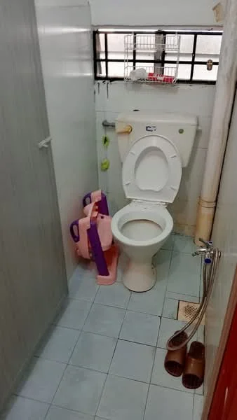 Bathroom features: white toilet, single-sink vanity, grey tiled walls, light blue tiled flooring, shower hose, plastic pink watering can, plastic purple bucket, window with metal grilles, wall-mounted soap dish, drain in floor.