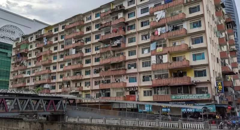 Exterior type: low-rise apartment block; features: multi-storey building, balconies with railings, exposed concrete facade, various laundry hanging on balconies, ground-level retail spaces, signage for restaurants, windows with metal grilles, painted facade in beige and orange, visible urban surroundings, adjacent to a bridge; style: traditional.