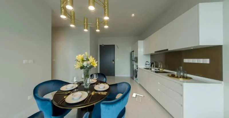 Dining features: round dining table, 4 upholstered dining chairs in blue, gold pendant lighting with multiple bulbs, white cabinetry, stainless-steel fridge, built-in oven, brown backsplash, white countertop, glass vase with flowers, table setting with plates and cutlery; style: modern. Kitchen features: white cabinetry, stainless-steel fridge, built-in oven, brown backsplash, white countertop; style: modern.