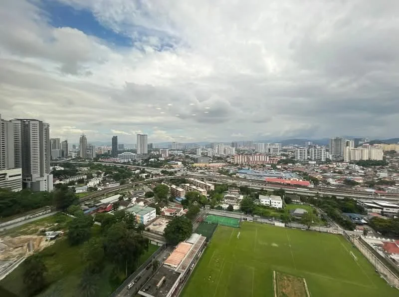 Exterior type: high-rise condominium; features: city skyline view, surrounded by greenery, sports field visible, urban landscape, multiple high-rise buildings, railway tracks visible, cloudy sky, open space in foreground; style: modern.