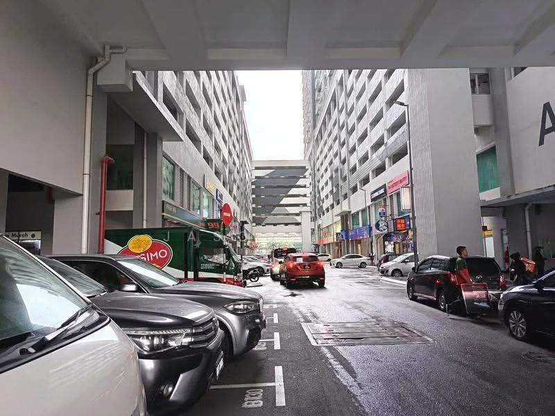 Exterior type: multi-storey car park; features: covered parking area, multiple parked vehicles, concrete structure, retail shops visible, rainy weather, wide driveway, signage on buildings, grey painted facade; style: modern.