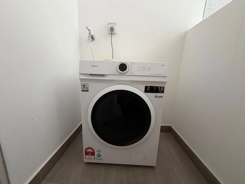 Laundry features: front-load washing machine, white appliance, 7 kg capacity, control panel with digital display, water inlet hose, drainage outlet, grey tile flooring, white walls.