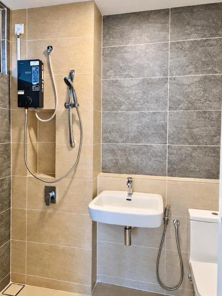 Bathroom features: beige tiled walls, grey textured tile accent wall, large-format wall tiles, light grey floor tiles, wall-mounted sink, chrome basin mixer, wall-mounted shower heater, handheld shower, shower hose, wall-mounted shower control, white toilet, compact bathroom layout, recessed shower area, chrome plumbing fixtures; style: modern.