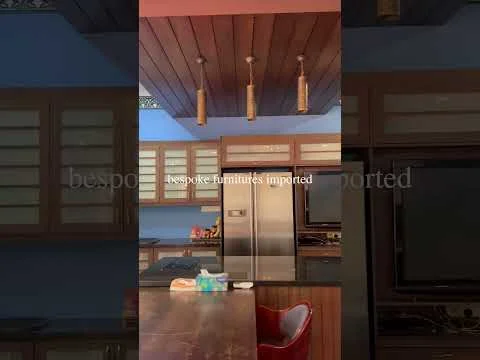 Kitchen features: wooden ceiling with pendant lights, brown cabinetry with glass inserts, stainless-steel fridge, kitchen countertop, built-in oven, microwave, kitchen island, red dining chair; style: modern.
