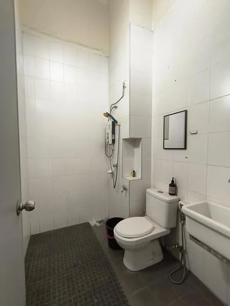 Bathroom features: white tiled walls, grey tiled flooring, one-piece toilet, shower area with handheld shower, mirror above sink, sink with cabinet, wall-mounted water heater, drainage channel, bucket, soap dispenser.