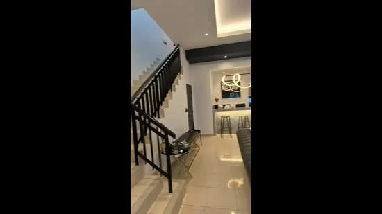 Living features: white painted walls, polished tile flooring, staircase with black railing, recessed downlights, chandelier, open-plan layout, bar counter with stools, kitchen area visible in background; style: modern.