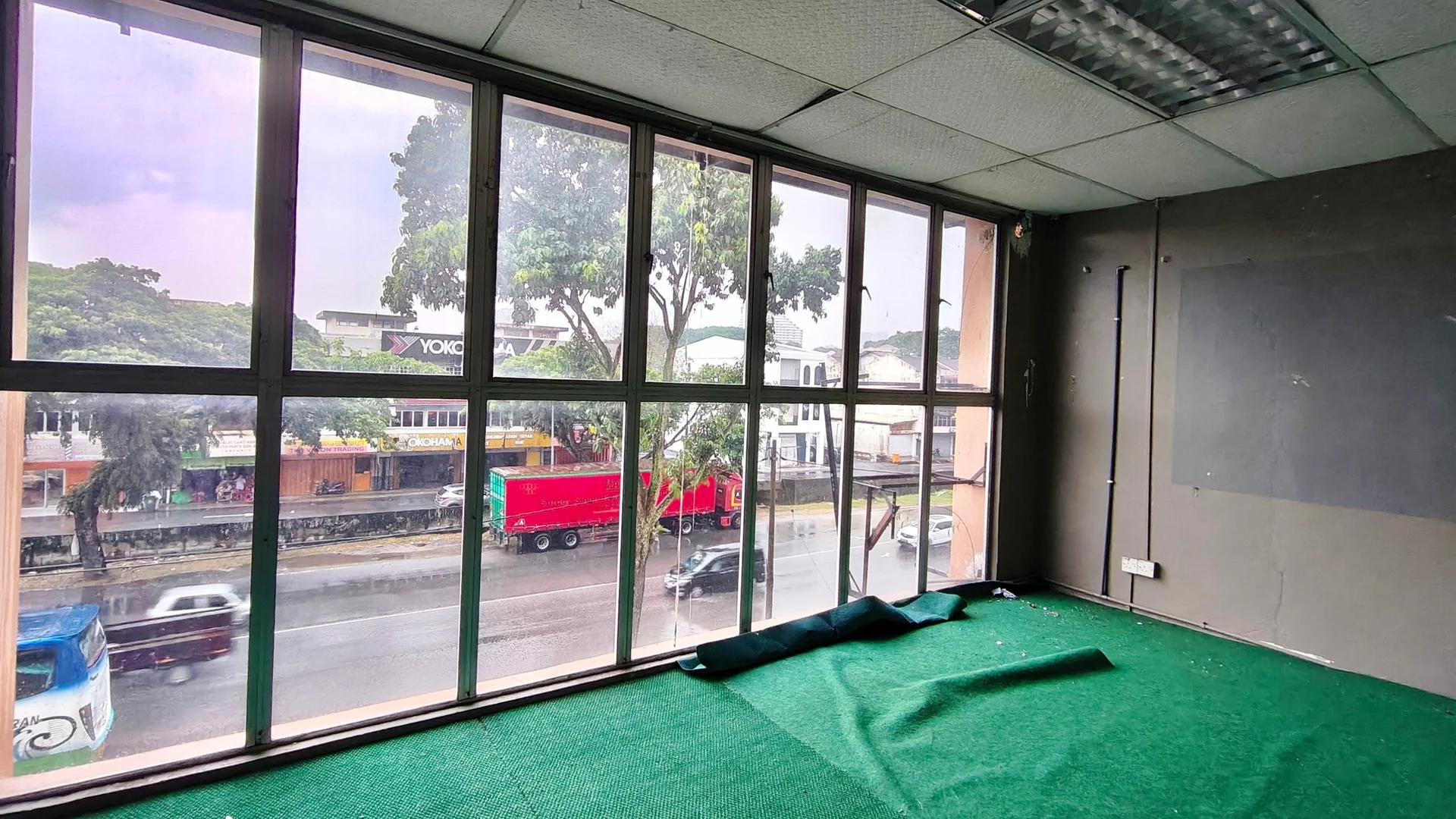 Room features: large floor-to-ceiling windows, green carpet flooring, grey painted walls, exposed ceiling tiles, visible electrical outlets, curtains or blinds, urban view, traffic visible outside; view: city.