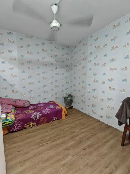 Bedroom features: wooden flooring, wallpaper with cartoon pattern, single bed with colorful bedding, ceiling fan, window, decorative throw pillows, small table fan, blanket, chair.