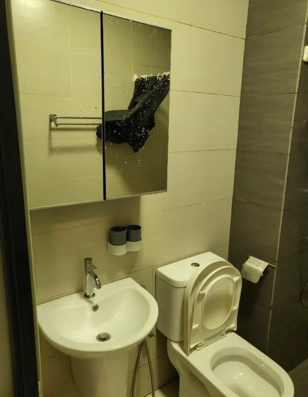Bathroom features: white ceramic sink, chrome basin mixer faucet, wall-mounted mirror with shelf, white toilet, toilet paper holder, grey tile wall, light-colored tile flooring, shower area with glass partition; style: modern.