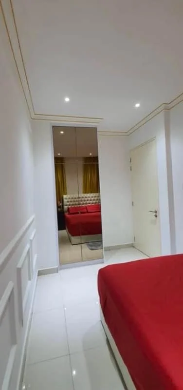 Bedroom features: red bedspread, white walls with crown moulding, mirror wardrobe, recessed downlights, white tiled flooring, curtains in yellow, double bed, door with handle; style: modern.