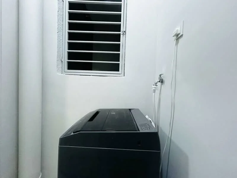 Laundry features: top-load washing machine in black, white painted walls, window with metal grille, water supply tap, drainage outlet.