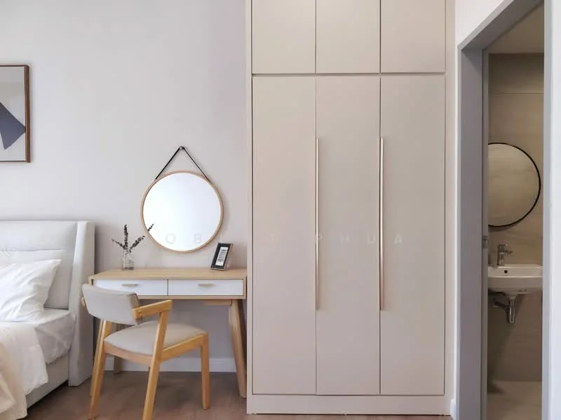 Room features: white walls, light wood flooring, built-in wardrobe with two doors, desk with two drawers, wooden chair, round mirror, small bathroom visible in background with circular mirror, neutral color palette; style: modern.
