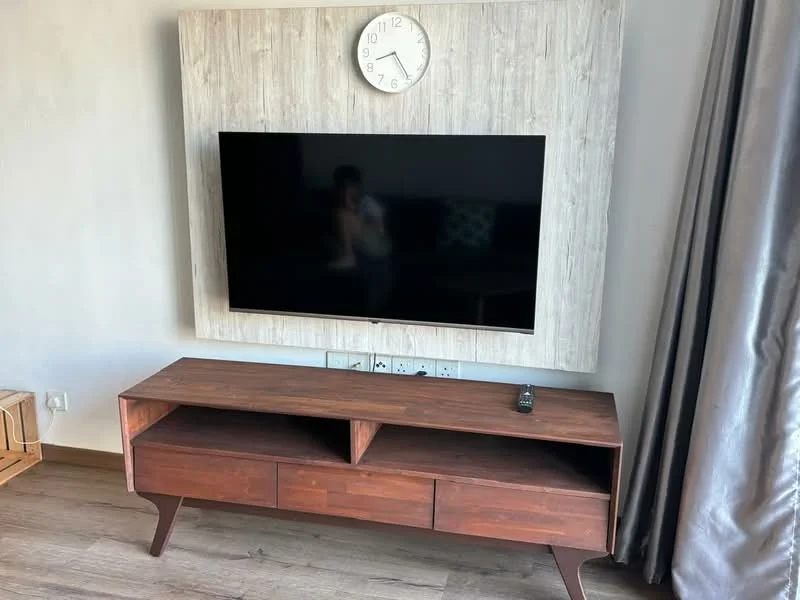 Living features: wall-mounted flat-screen TV, wooden TV console with drawers, light wood feature wall, clock on wall, laminate flooring, grey curtains, decorative cushions on sofa, neutral wall color; style: modern.