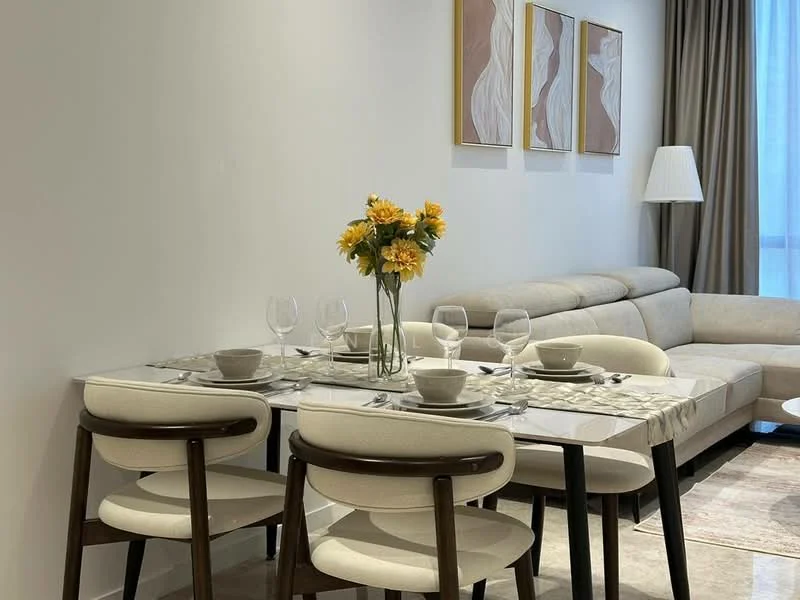Dining features: 6-seater dining table with marble top, upholstered dining chairs, decorative vase with yellow flowers, table setting with plates and glasses, neutral wall colour, soft lighting from nearby lamp, area rug under dining table, framed artwork on wall; style: modern. Living features: sectional sofa in light colour, floor lamp, curtains, neutral wall colour, decorative artwork on wall; style: modern.