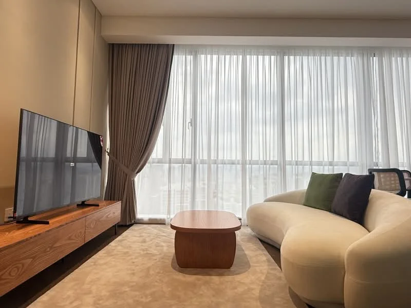 Living features: curved white fabric sofa, wooden coffee table, TV console with wooden finish, wall-mounted flat-screen TV, floor-to-ceiling windows, sheer curtains, beige carpet flooring, decorative cushions in green and black; style: modern; view: city.