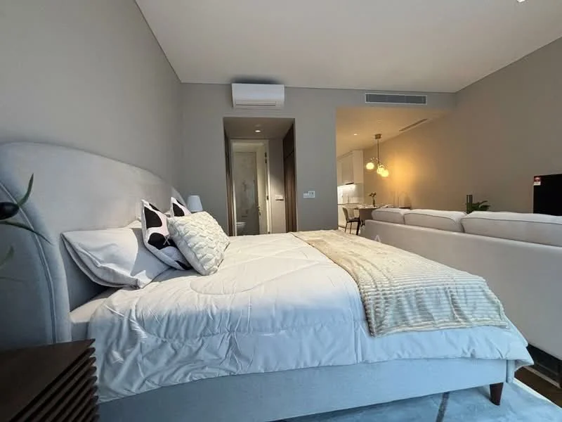 Bedroom features: king-size bed with upholstered headboard, white bedding, decorative pillows, nightstand with lamp, wall-mounted air conditioning unit, grey painted walls, carpet flooring, floor-to-ceiling windows, sofa in the background, open-plan layout to living area; style: modern.