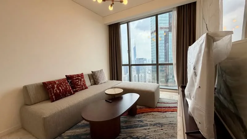 Living features: large floor-to-ceiling windows, grey fabric sectional sofa, decorative cushions in red and white, round wooden coffee table, area rug with geometric pattern, wall-mounted TV, wooden TV console, curtains, marble flooring, city skyline view; style: modern; view: city.