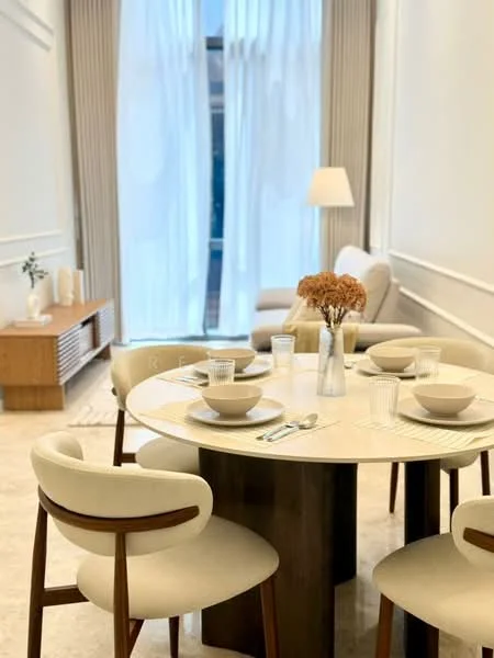 Dining features: round dining table with marble top, 4 upholstered dining chairs in cream, decorative vase with dried flowers, table setting with bowls and cutlery, light-colored wall with wainscoting, large window with sheer curtains, wooden sideboard, flooring with light-colored tiles, soft ambient lighting from lamp, sofa in background; style: modern.