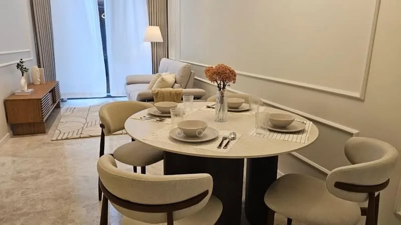 Dining features: round dining table, 4 dining chairs with upholstered seats, table setting with plates and glasses, decorative vase with dried flowers, area rug with geometric pattern, light-colored walls, neutral-colored flooring, soft lighting from floor lamp, sofa in adjacent living area, wooden sideboard, curtains covering large window; style: modern.