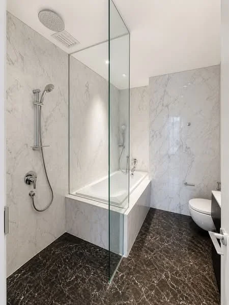 Bathroom features: marble wall cladding, glass shower enclosure, bathtub, chrome shower head, handheld shower, wall-mounted faucet, one-piece toilet, dark marble flooring, built-in vanity with storage, recessed lighting; style: modern.