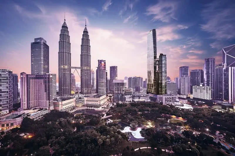 Exterior type: high-rise condominium; features: glass curtain wall, surrounded by mature greenery, urban skyline view, landscape garden, park area, multiple high-rise buildings, Petronas Twin Towers in background; style: modern.