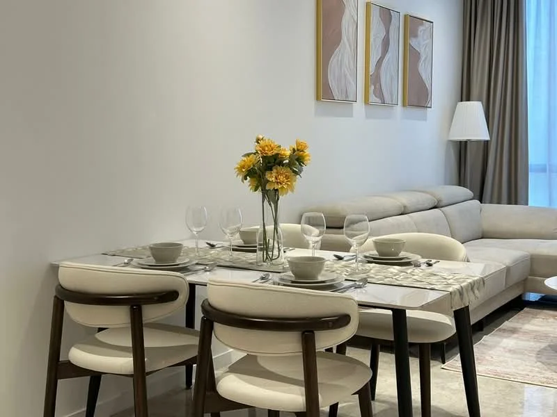 Dining features: marble dining table, 4 upholstered dining chairs, decorative vase with flowers, set of dinner plates, set of wine glasses, table placemats, neutral wall colour, soft lighting from lamp, beige sectional sofa in background, curtains, artwork on wall; style: modern.