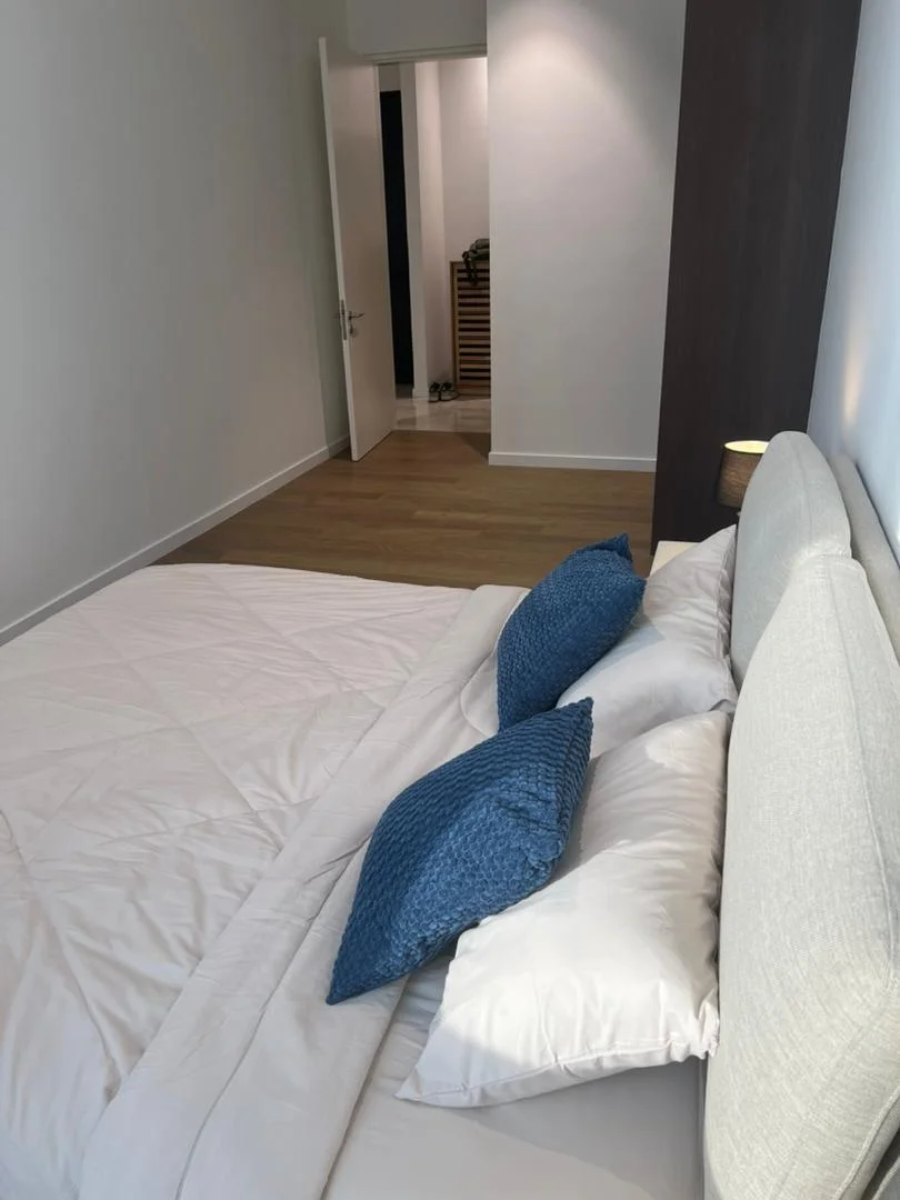Bedroom features: queen-size bed with upholstered headboard, white bedding, blue decorative pillows, wooden flooring, wall-mounted bedside lamp, neutral painted walls, open door leading to hallway, minimalist decor; style: modern.