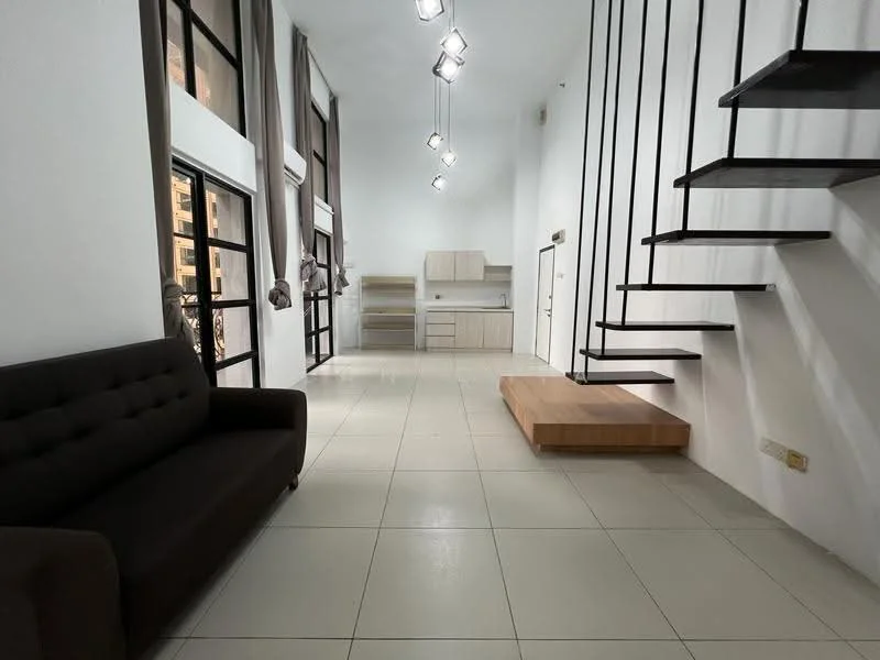 Living features: grey fabric sofa, white walls, tile flooring, recessed LED downlights, large windows with black frames, open layout, wooden platform, metal staircase, kitchen area visible in background, curtains, high ceiling; style: modern.
