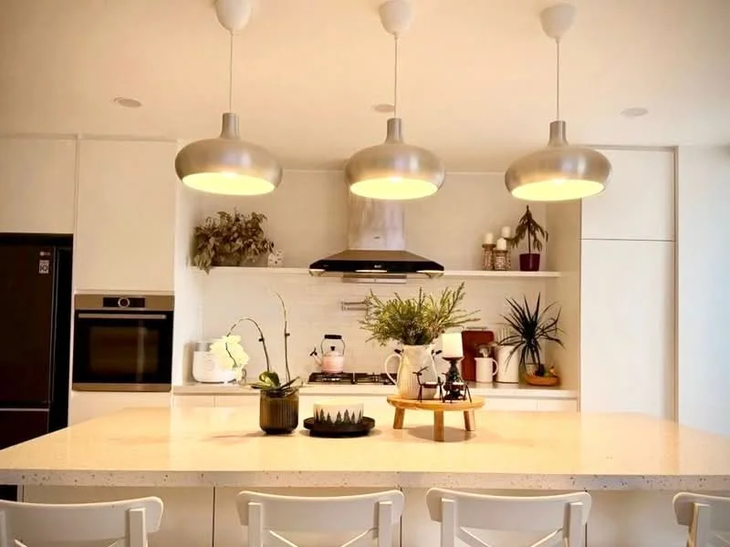 Kitchen features: white cabinetry, stainless-steel appliances, built-in oven, 4-burner gas hob, stainless-steel range hood, quartz countertop, pendant lighting with metallic finish, open shelving, decorative plants, kitchen island with seating, ceramic decorative items; style: modern.