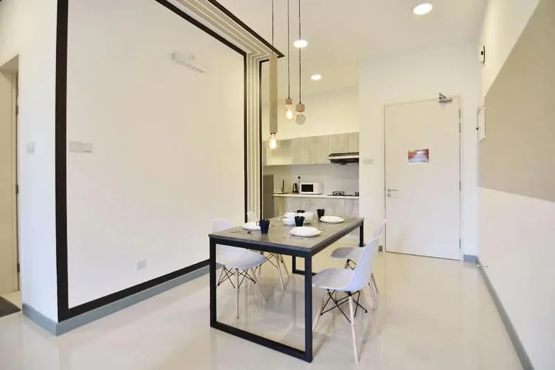 Dining features: dining table with black metal frame, 4 white plastic dining chairs, pendant lighting with exposed bulbs, white walls, grey accent wall, kitchenette visible in background, built-in kitchen cabinets, stainless-steel sink, countertop with appliances, recessed ceiling lights, flooring with glossy finish; style: modern.