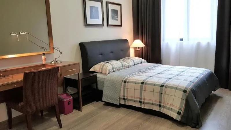 Bedroom features: queen-size bed with plaid bedding, upholstered headboard, wooden bedside lamp, wooden desk with lamp, framed artwork on walls, large window with sheer curtains, wooden flooring, brown woven chair, small pink storage box; style: modern.