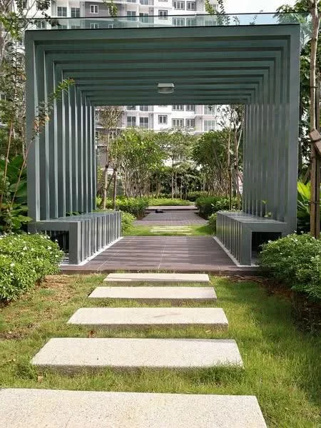 Exterior type: landscape garden; features: stepping stone pathway, green manicured lawn, landscaped garden with shrubs, metal archway structure, benches on either side of pathway, surrounding greenery, paved walkway, residential buildings in background; style: modern.