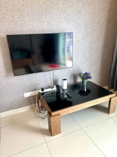 Living features: flat-screen TV mounted on wall, black glass coffee table with wooden legs, decorative vase with flowers, white walls with textured finish, light-colored floor tiles, cable management visible, curtains in grey fabric; style: modern.