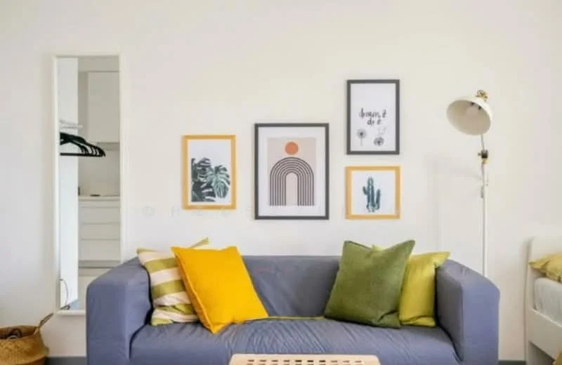 Living features: grey fabric sofa, yellow and green decorative cushions, framed artwork on walls, white painted walls, wooden coffee table, floor lamp, open closet with hangers visible, light-colored flooring; style: modern.