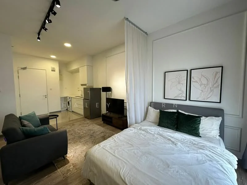 Room features: queen-size bed with white bedding, grey upholstered headboard, 2 green decorative pillows, sofa in dark grey fabric, coffee table, wall-mounted TV, wooden flooring, recessed downlights, white walls, framed artwork on walls, kitchen area with white cabinetry, stainless-steel fridge, open layout; style: modern.