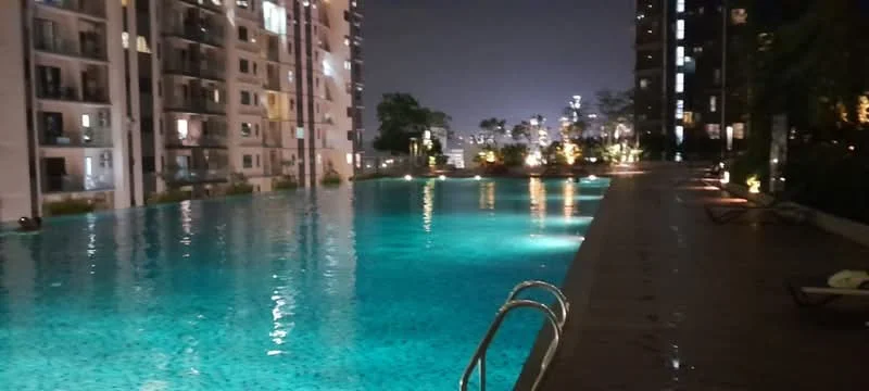Exterior type: swimming pool deck; features: infinity pool, pool deck with loungers, landscaped garden area, night lighting, surrounding residential buildings, city skyline in background; style: modern.
